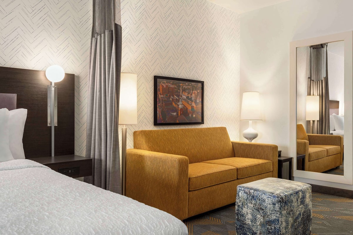 A hotel room with a large bed, a sofa, a small ottoman, a nightstand, a lamp, a mirror, and a framed picture on the wall.