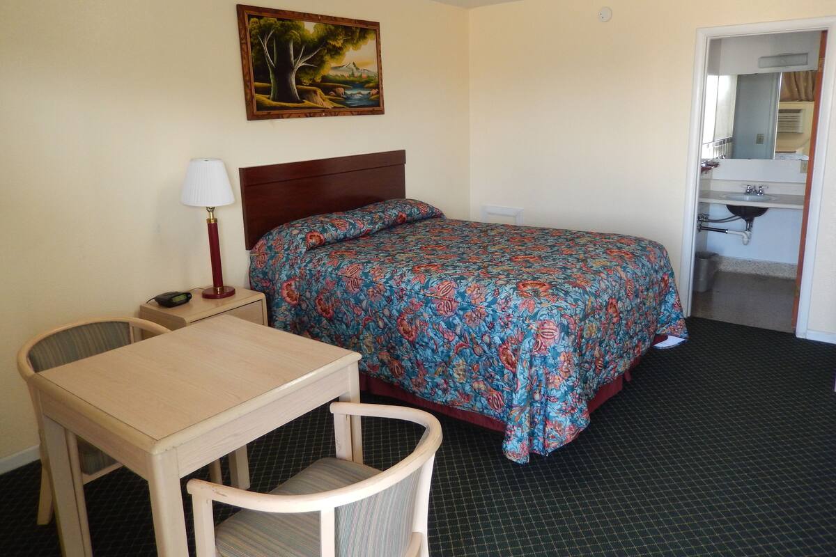 A hotel room with a bed, a desk, a chair, a lamp, and a painting on the wall.