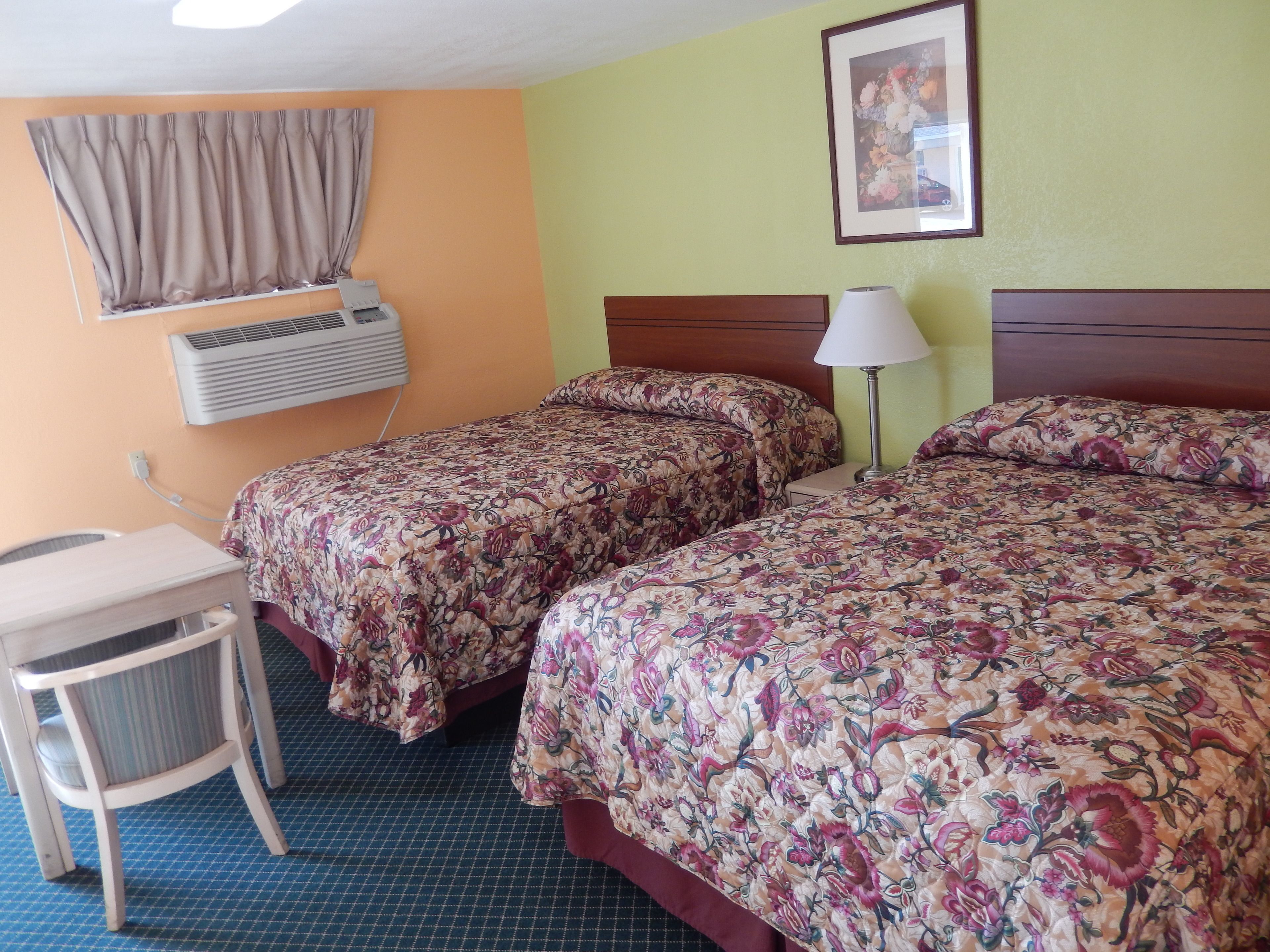 A hotel room with two beds, a desk, a chair, a lamp, an air conditioning unit, and a framed picture on the wall.