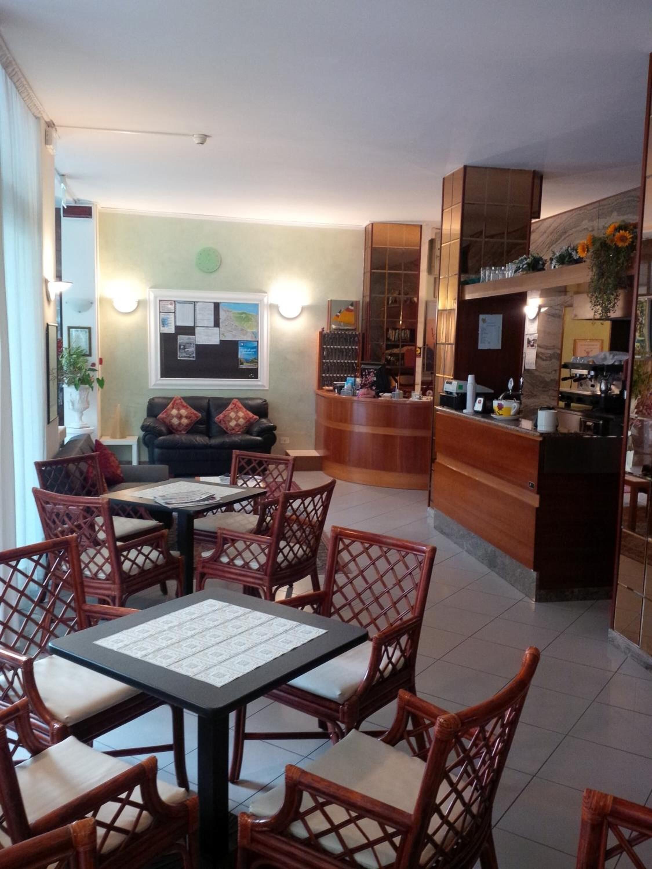 A hotel lobby with a reception desk, a seating area with wicker chairs and a table, a sofa, and a bulletin board with notices.