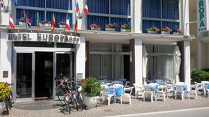 A hotel with a glass entrance, outdoor seating area with tables and chairs, and multiple flags displayed outside. - Hotel Europa (Gabicce Mare)