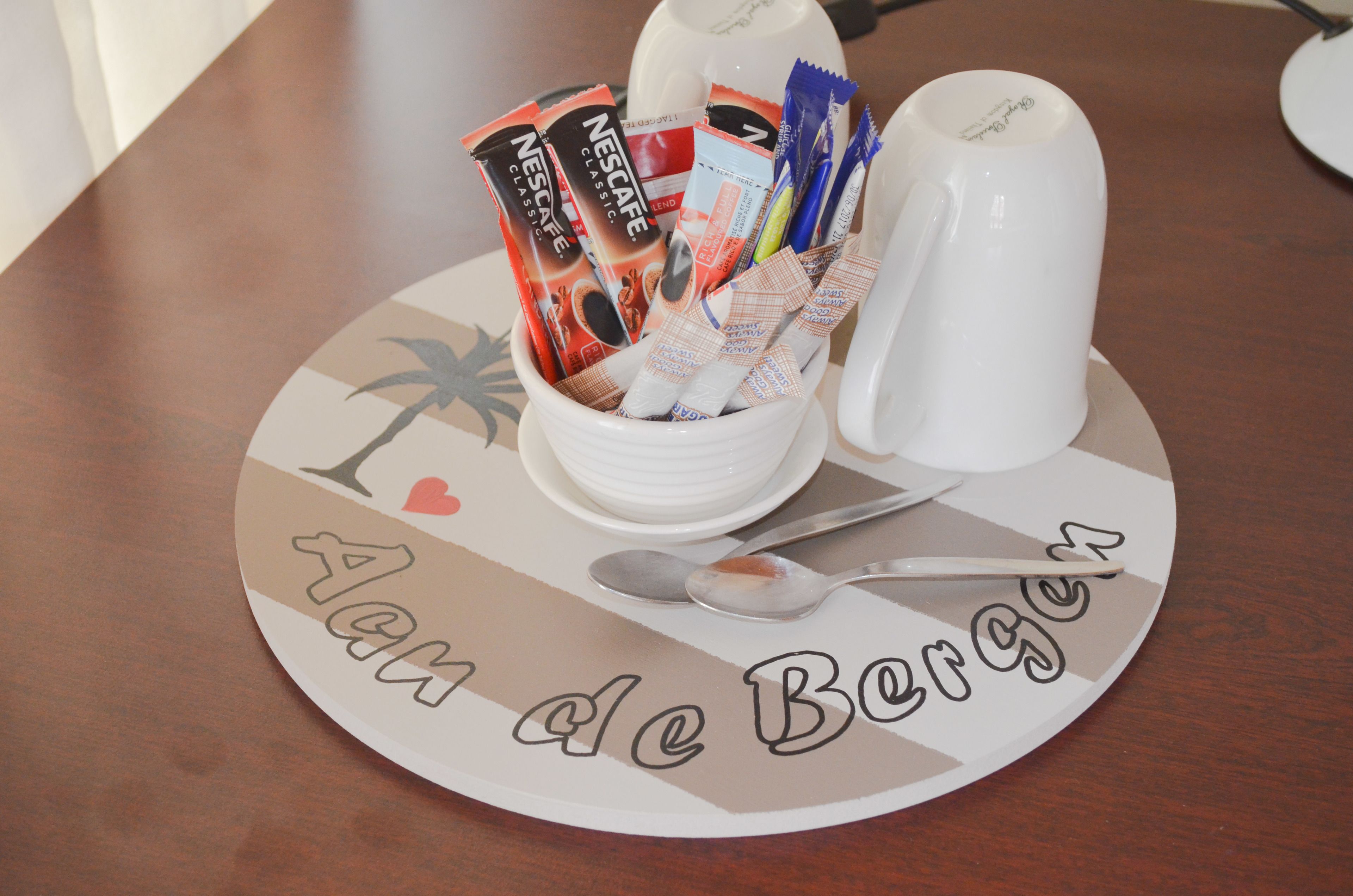 A breakfast setup with a coffee maker, a cup, and a bowl of assorted coffee packets on a placemat with the text "Aline de Bergue".