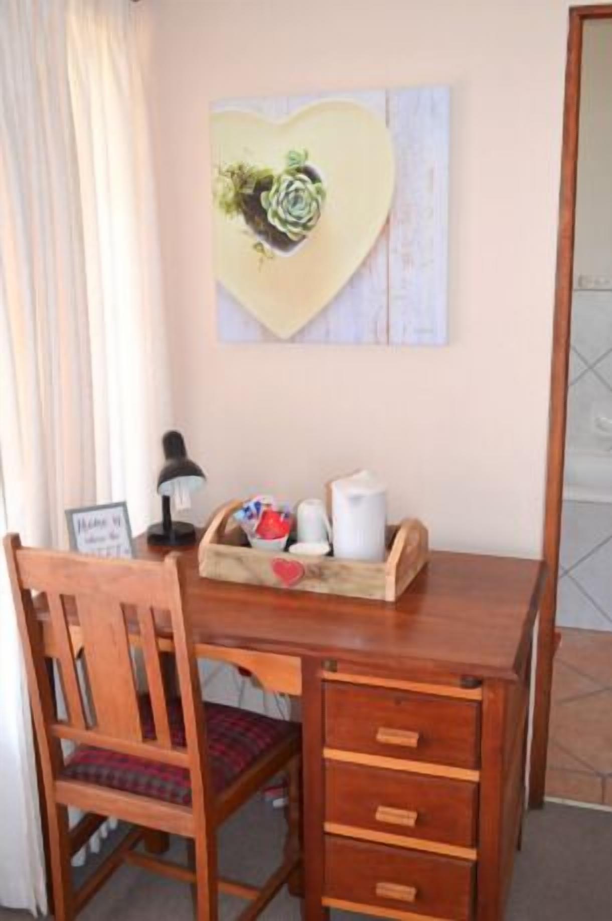A wooden desk with a chair, a heart-shaped artwork, a lamp, and a tray with cups and a pitcher.