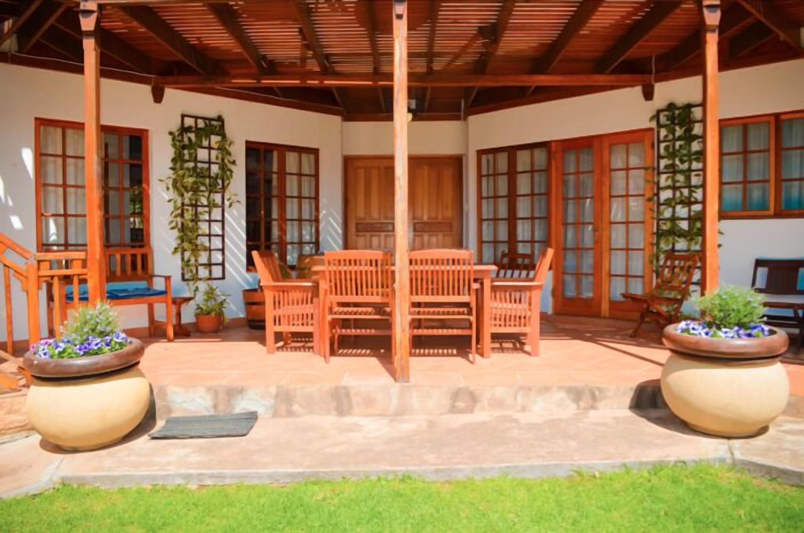 A covered patio area with a wooden table and chairs, surrounded by potted plants and a wooden fence.
