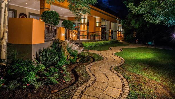 A hotel with a curved pathway leading to a gated entrance, surrounded by lush greenery and a well-maintained lawn. - Home Guesthouse (Rustenburg)