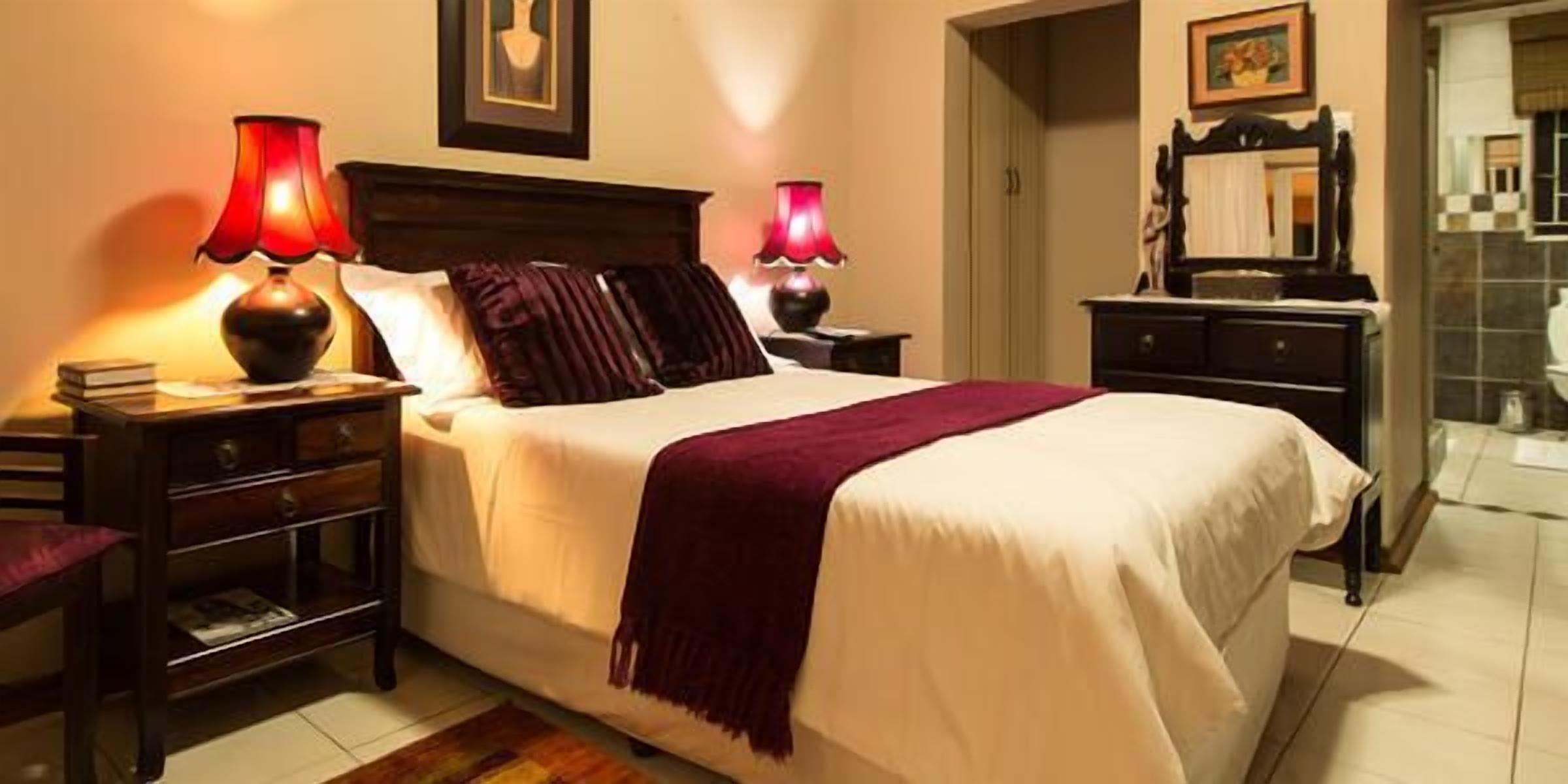 A neatly made bed with a white and maroon bedspread, two bedside tables with lamps, a wooden nightstand, and a dresser with a mirror.