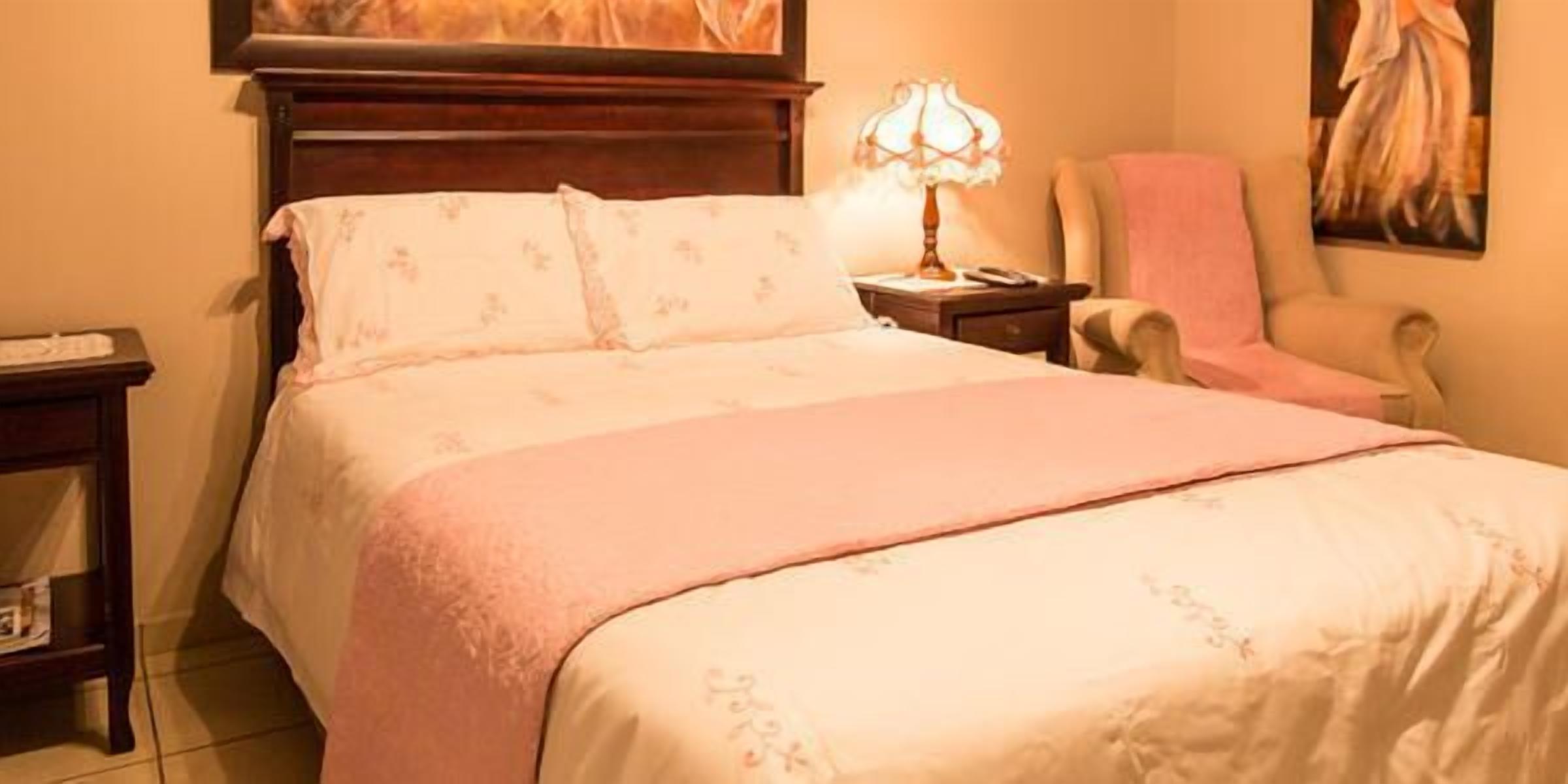 A neatly made bed with a pink blanket and floral bedding, a bedside lamp, a chair, and a painting on the wall.