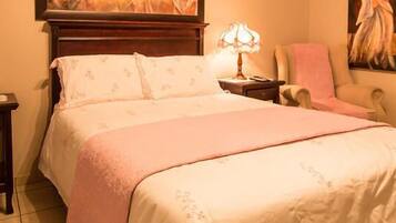 A neatly made bed with a pink blanket and floral bedding, a bedside lamp, a chair, and a painting on the wall.
