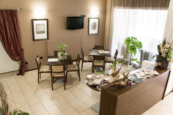 A hotel room with a dining area, a TV mounted on the wall, and a reception desk with magazines and a plant. - Hotel Fontanelle (Bertinoro)