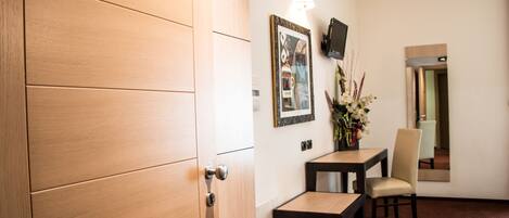 A hotel room with a wooden door, a small table with a chair, a wall-mounted TV, and a framed picture.