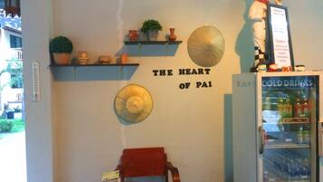 A restaurant interior with a wall-mounted sign reading "THE HEART OF PAI," a small table with a stack of books, a red chair, and a refrigerator labeled "COLD DRINKS."