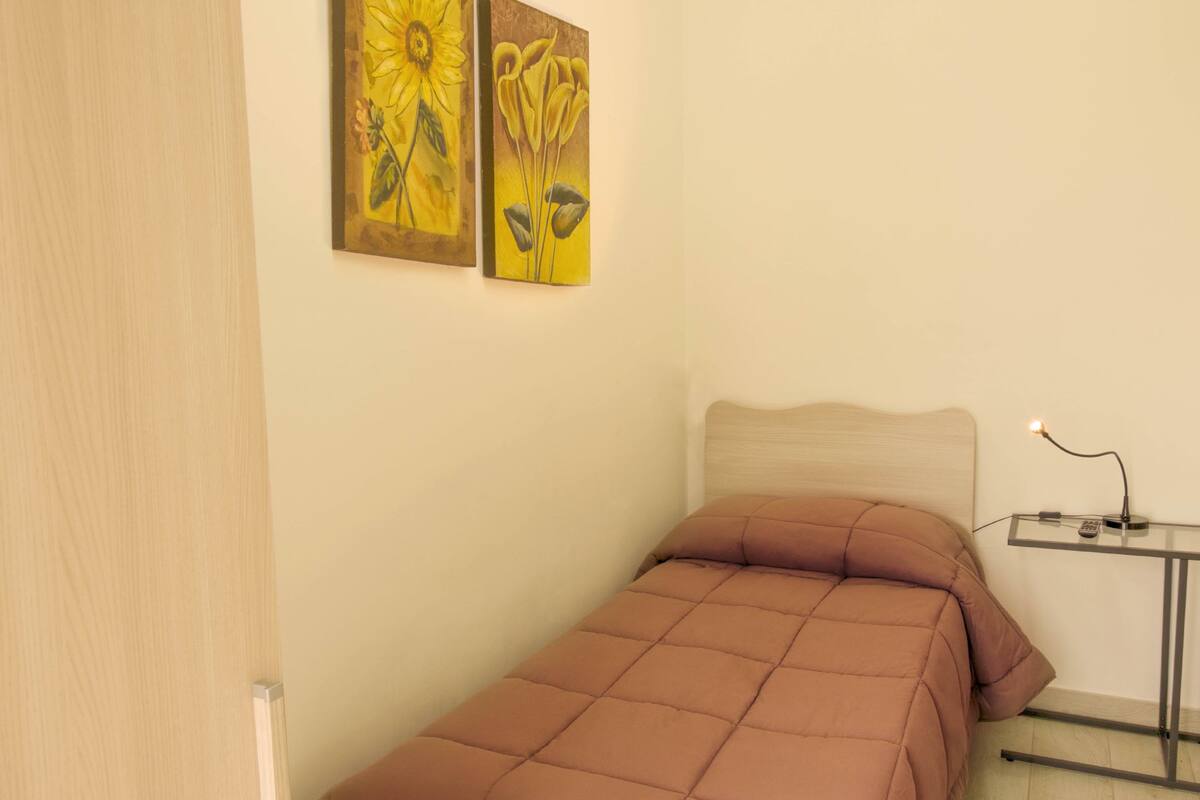 Una cama pequeña y ordenada con cabecera, una mesita de noche con lámpara, y dos cuadros en la pared.