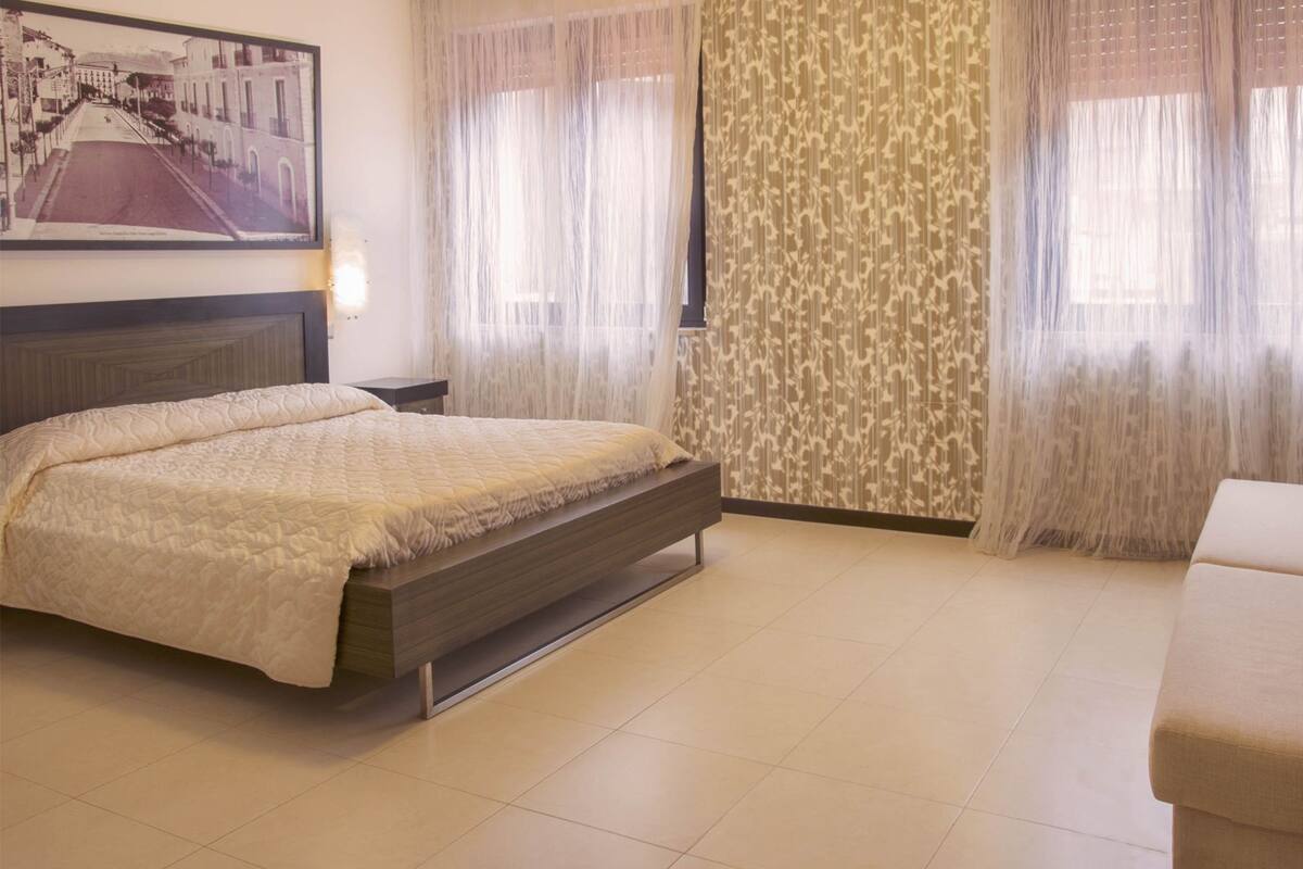 Un dormitorio con cama, mesita de noche, lámpara de pared, ventana con cortinas de gasa y un papel tapiz estampado.
