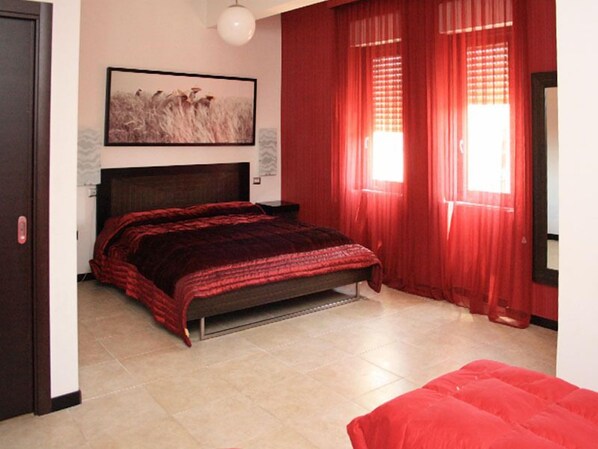 Un dormitorio con cama, cabecera, ventana con cortinas rojas y un cuadro colgado en la pared.