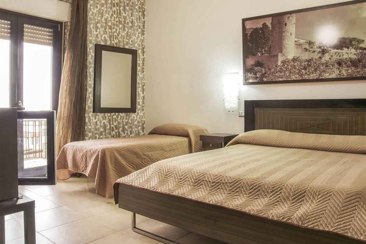 Habitación de hotel con dos camas individuales, un espejo grande, un cuadro de un castillo y vistas a un balcón.
