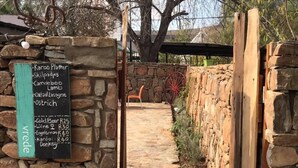 A stone pathway leading to a rustic outdoor seating area with a menu board listing food and drink prices. - The Ibis Lounge (Nieu-Bethesda)