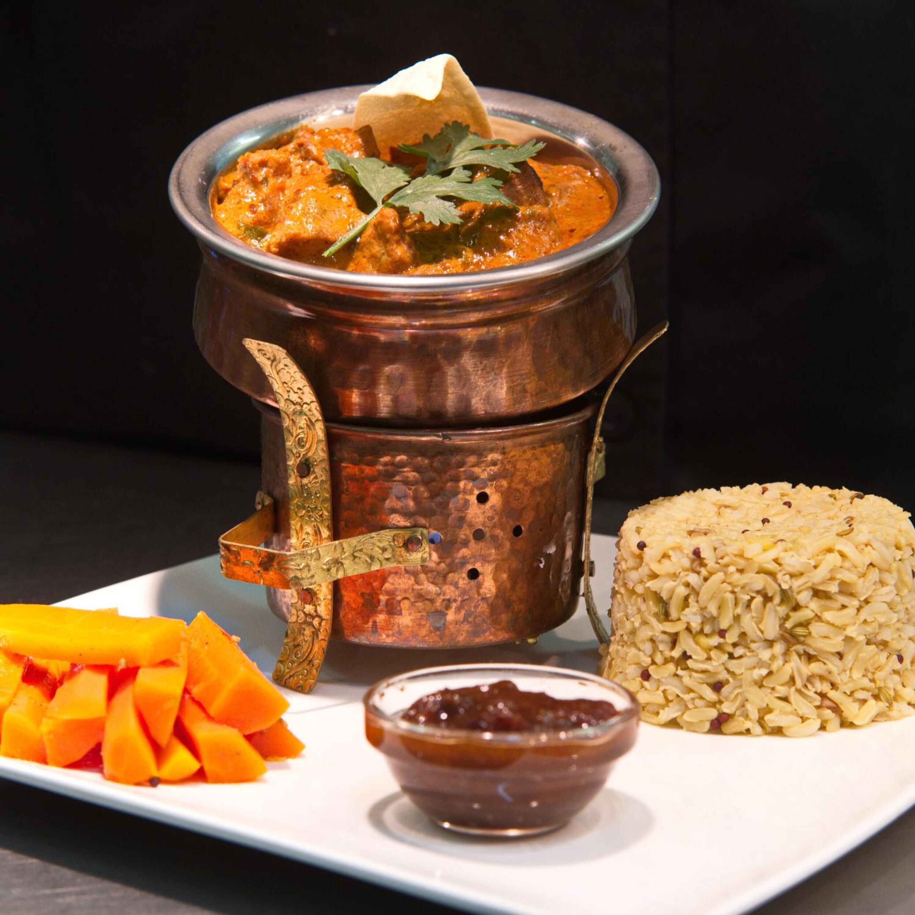 A traditional copper pot with a lid, accompanied by a plate of rice, mango slices, and a small bowl of chutney.