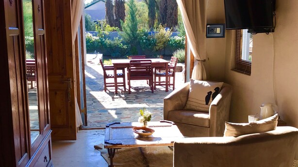 A room with a wooden desk, a leather sofa, a television mounted on the wall, and a view of the garden through a large window. - The Ibis Lounge (Nieu-Bethesda)