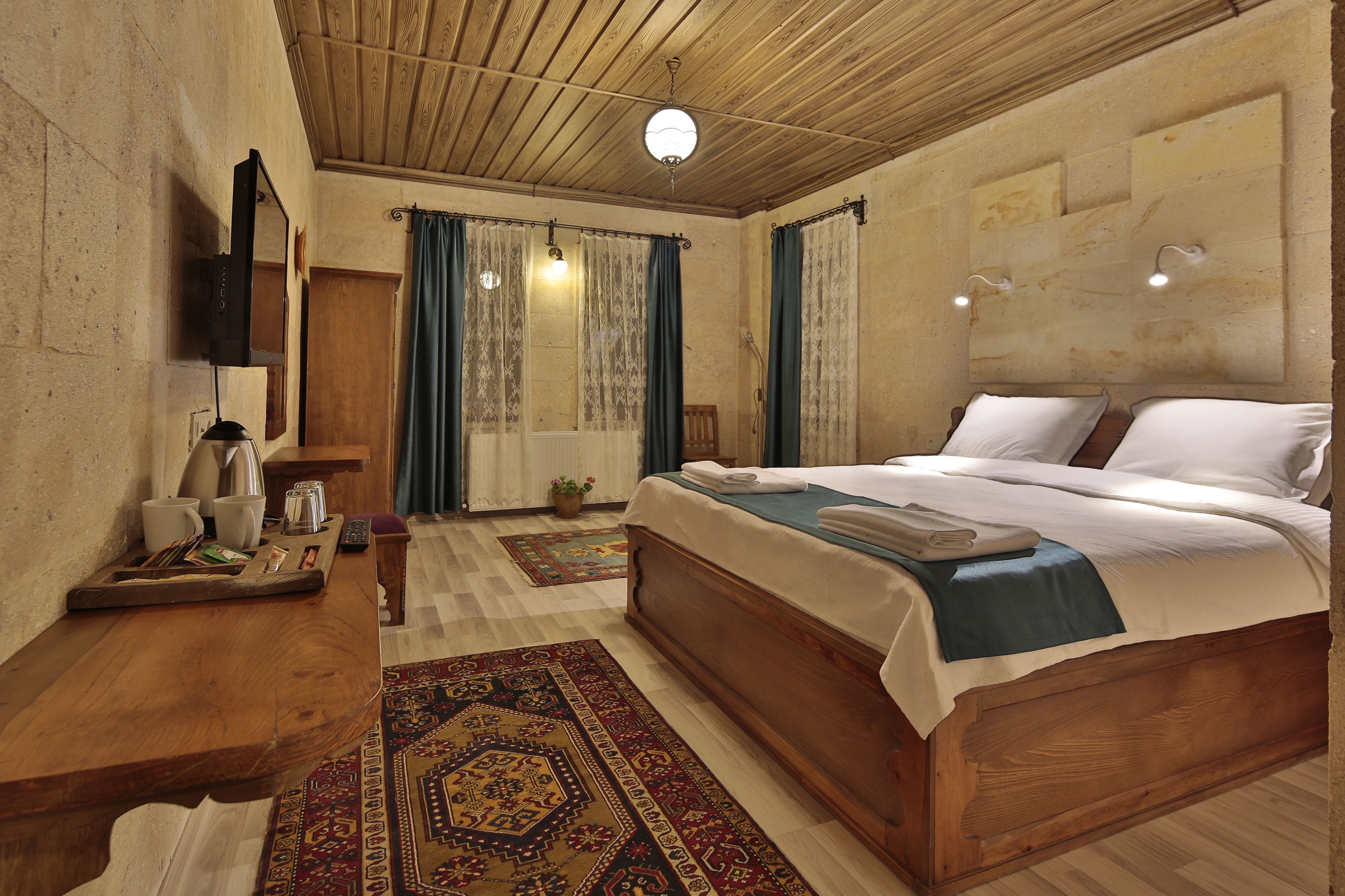 A bedroom with a wooden bed, a desk with a kettle, and a TV on the wall.