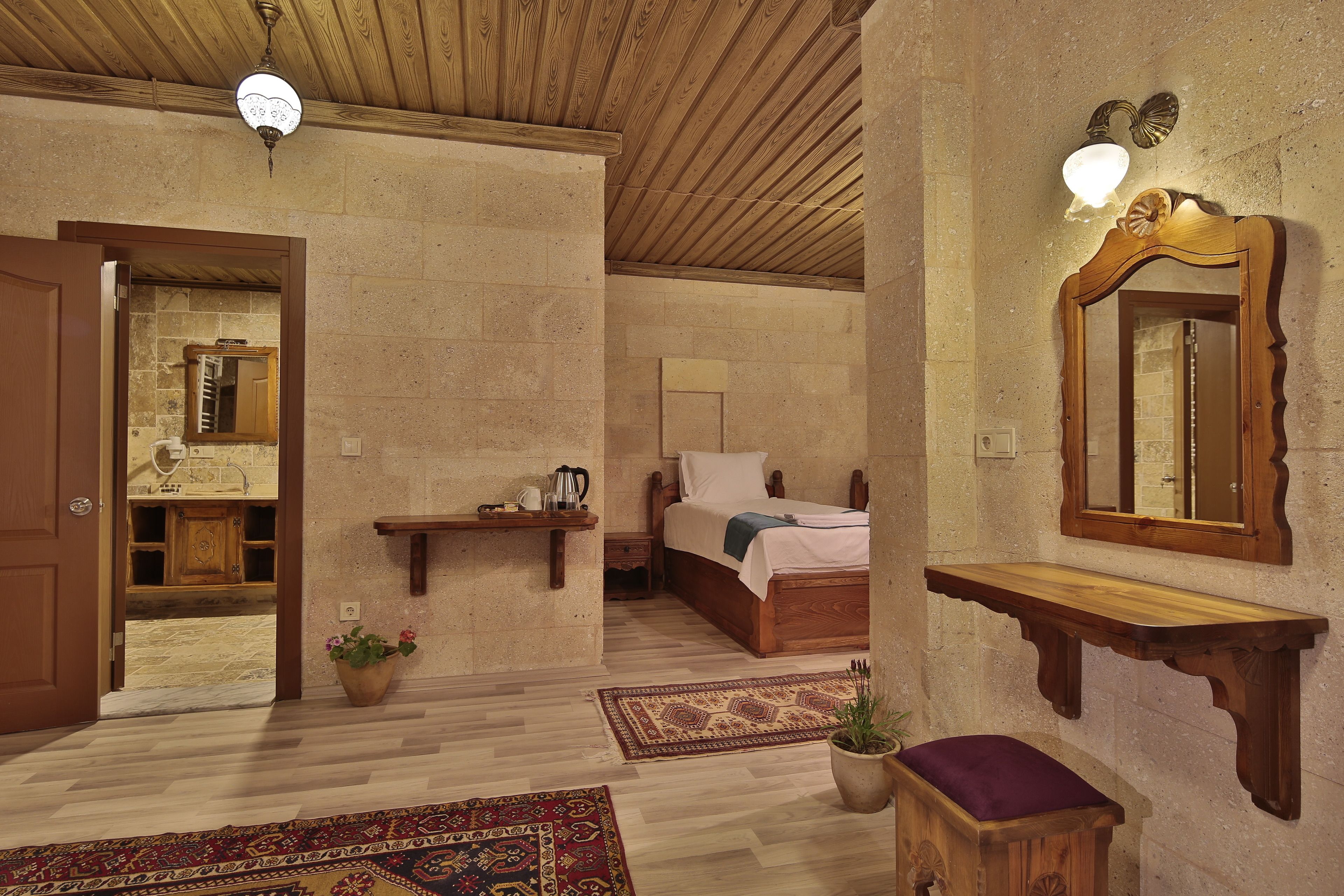 A room with a wooden ceiling, a bed with a white mattress and a wooden headboard, a wooden bench, a wall-mounted mirror, and a small table with a lamp.