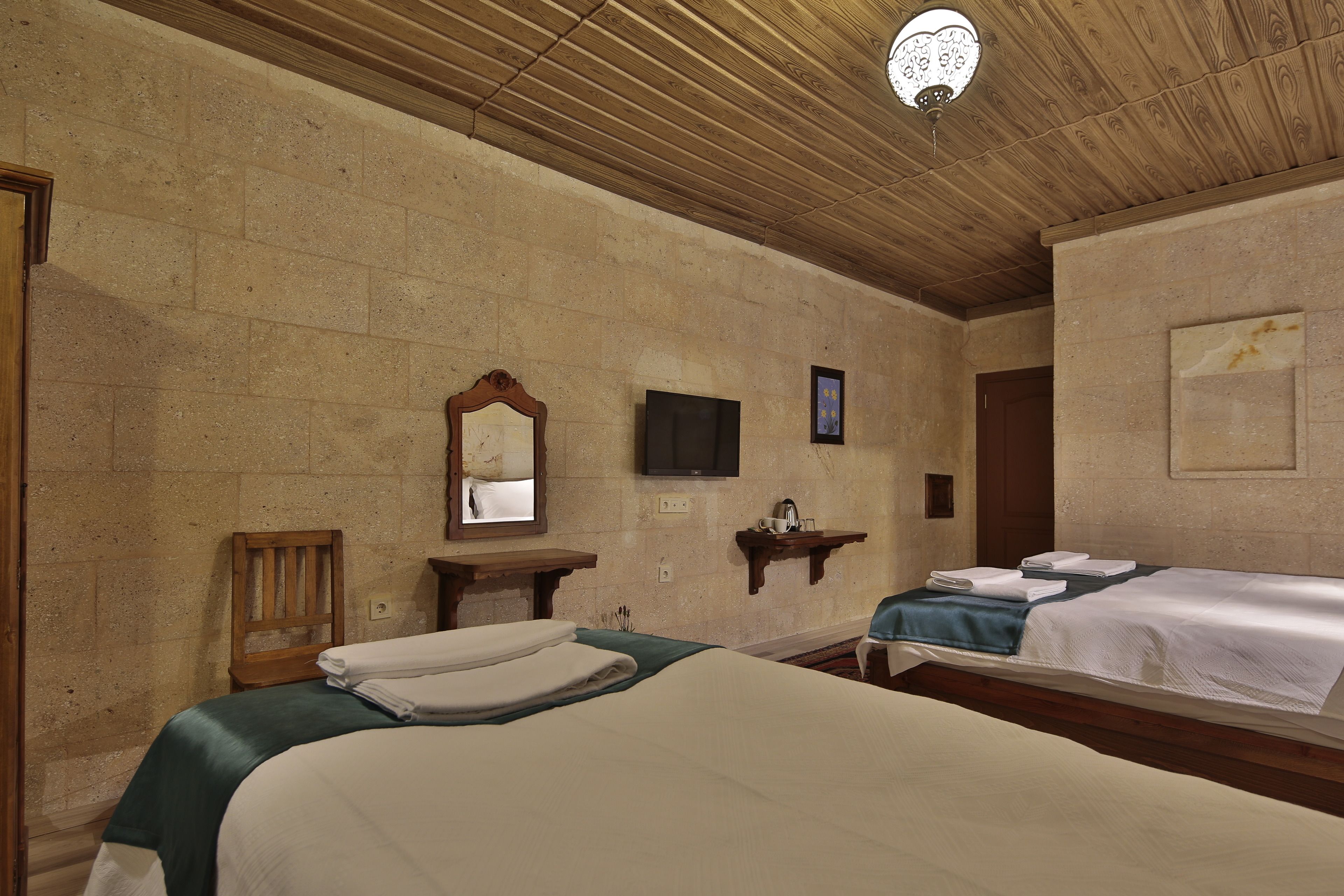 A hotel room with two beds, a wooden ceiling, stone walls, a flat-screen TV, and a small shelf with a cup and a book.