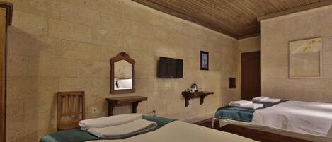 A hotel room with two beds, a wooden ceiling, stone walls, a flat-screen TV, and a small shelf with a cup and a book.