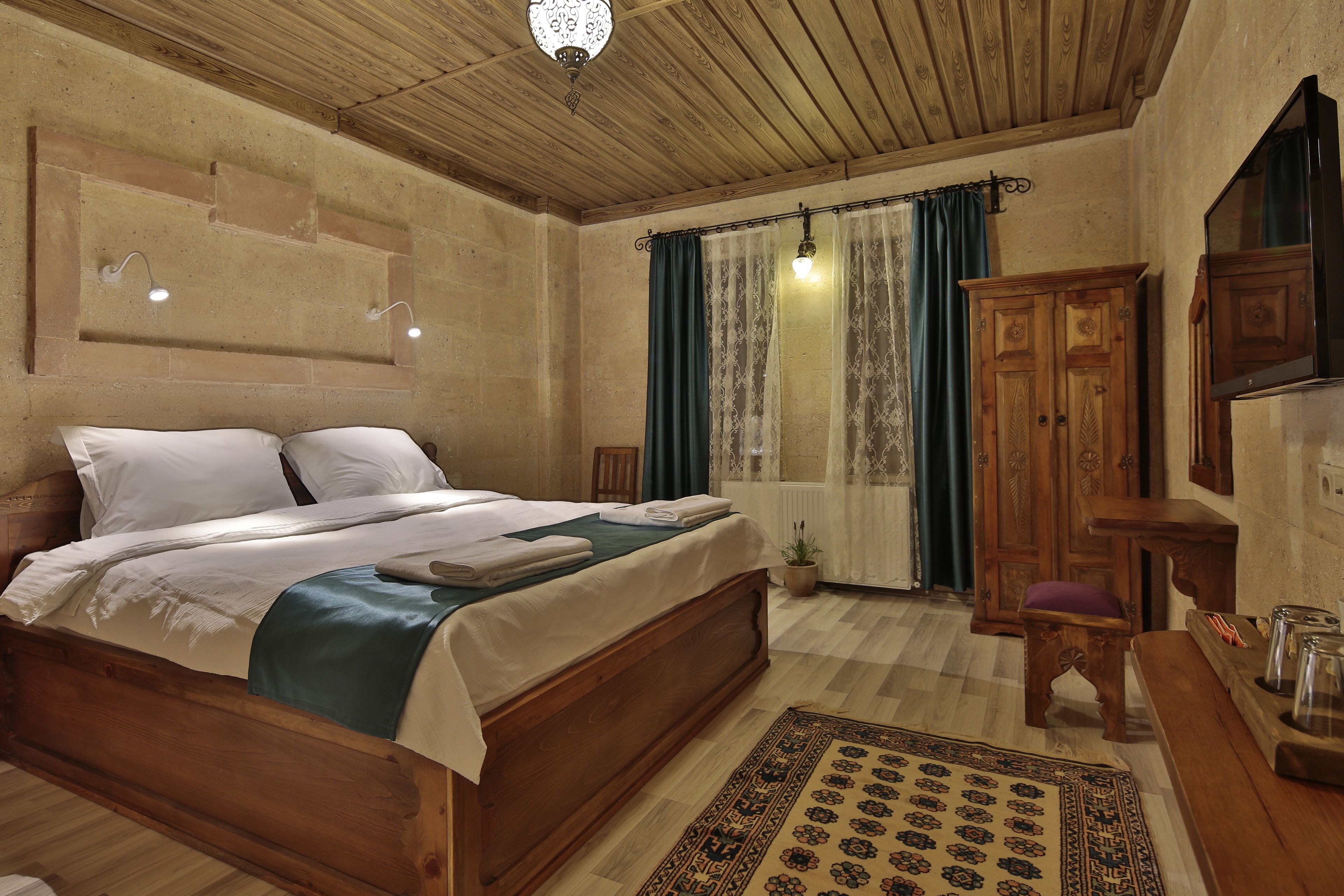 A wooden bed with white linens, a small wooden table with a vase, and a wooden wardrobe in a room with a checkered floor and a patterned rug.