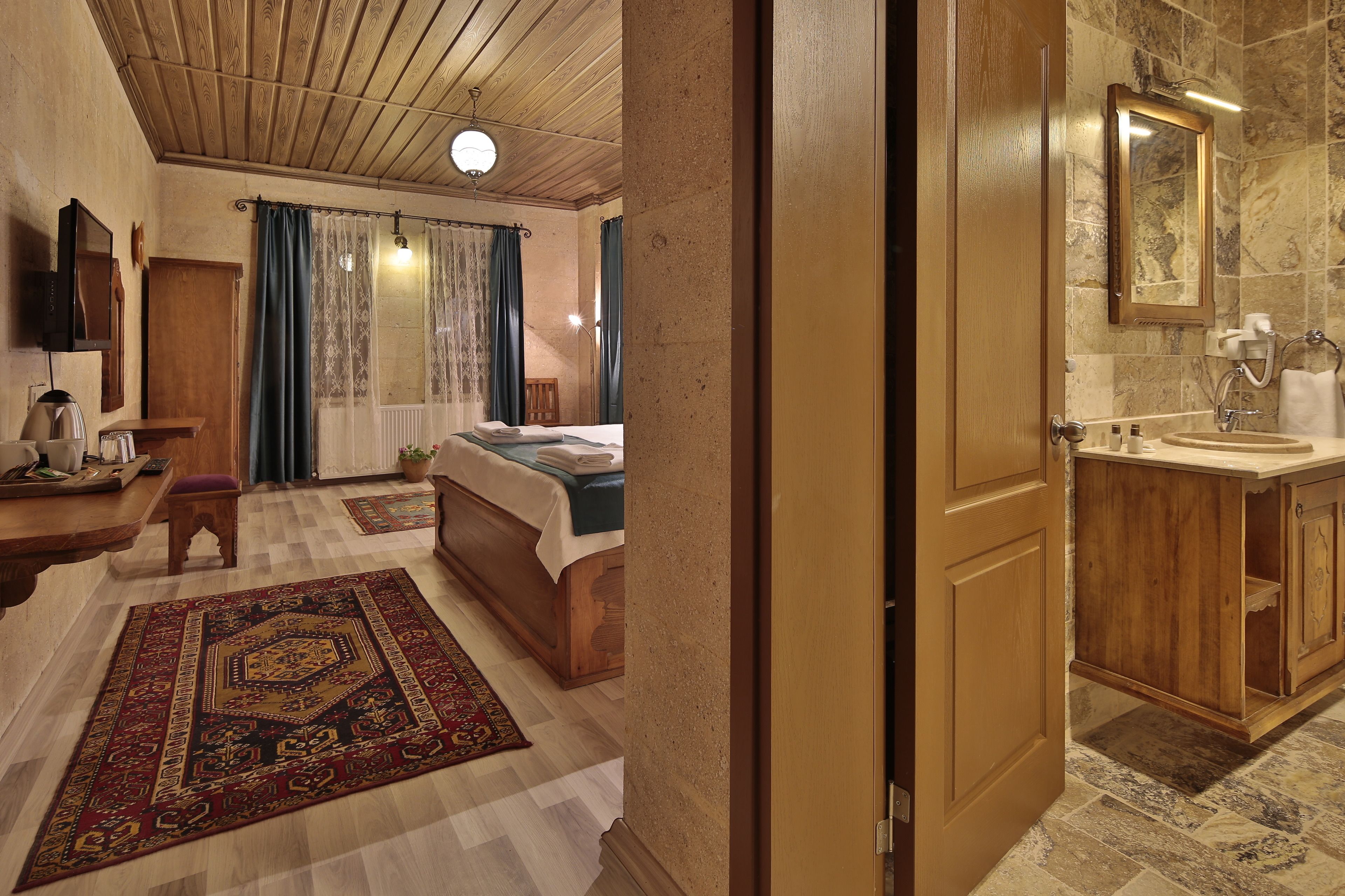 A hotel room with a wooden ceiling, a bed with a blue bedspread, a wooden desk with a lamp, a bathroom with a marble sink, and a large mirror.
