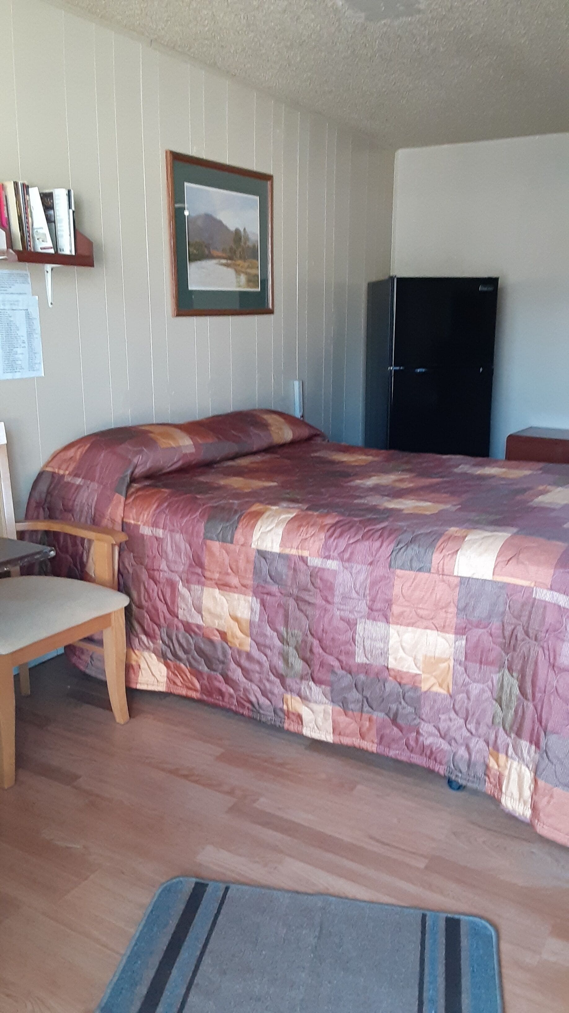 Un dormitorio con cama, una silla, un escritorio, un cuadro en la pared y un refrigerador.