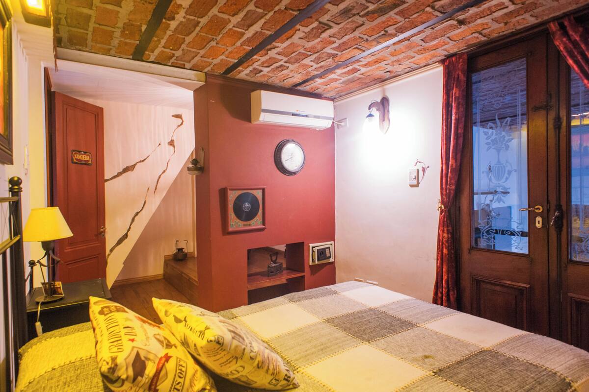 A room with a brick ceiling, a bed with a checkered bedspread, a clock on the wall, and a door with a window.