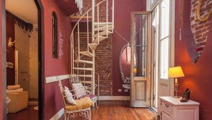 Una habitación con una escalera de caracol, una silla de metal blanco, suelo de madera y un armario blanco con una lámpara.