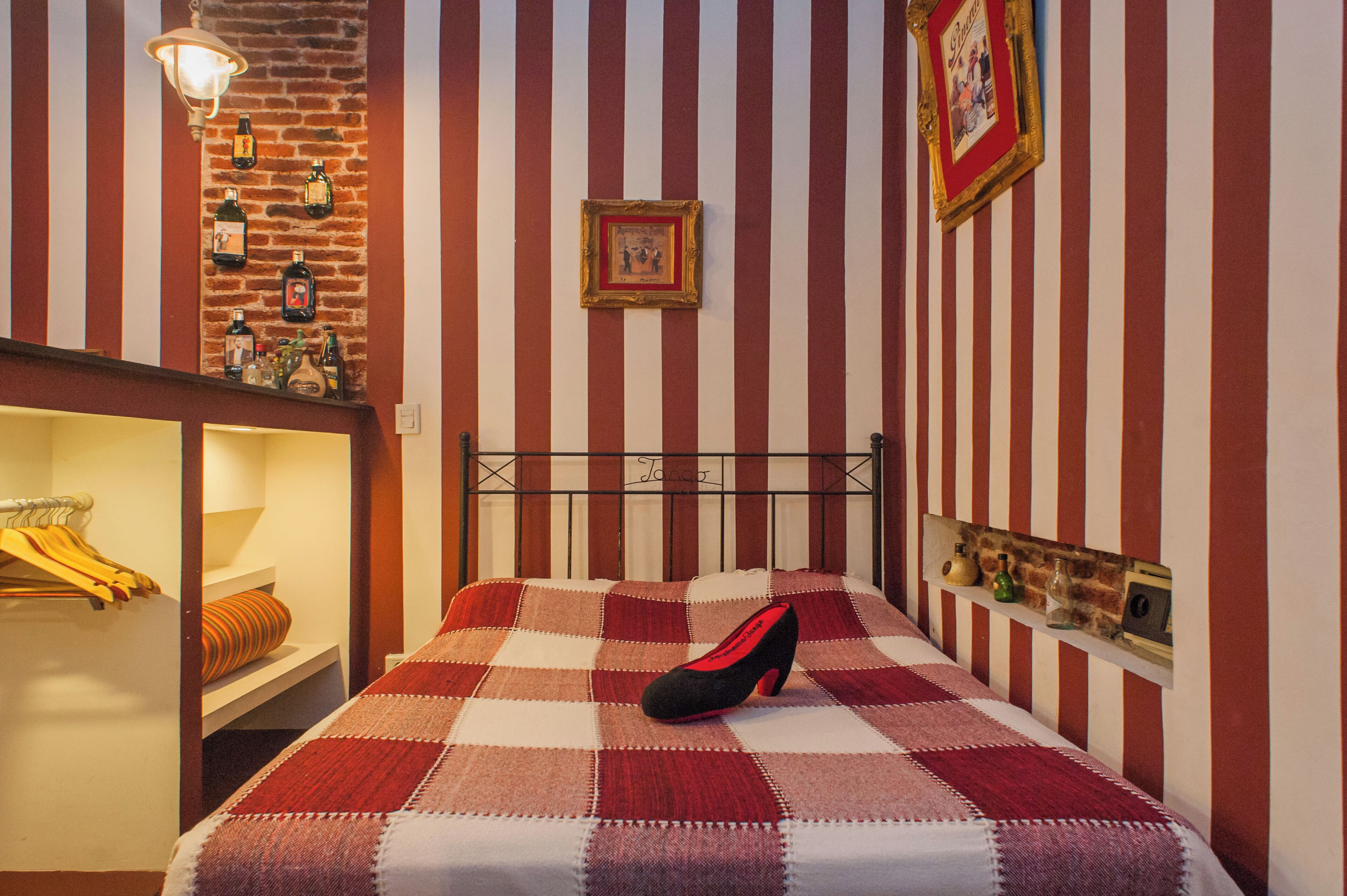 A bedroom with a checkered bedspread, a single black and red shoe on the bed, a brick wall, and a shelf with bottles and a picture frame.