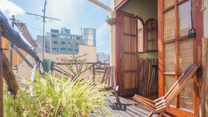 Una terraza de madera con una silla, una planta colgante y vistas de edificios al fondo.