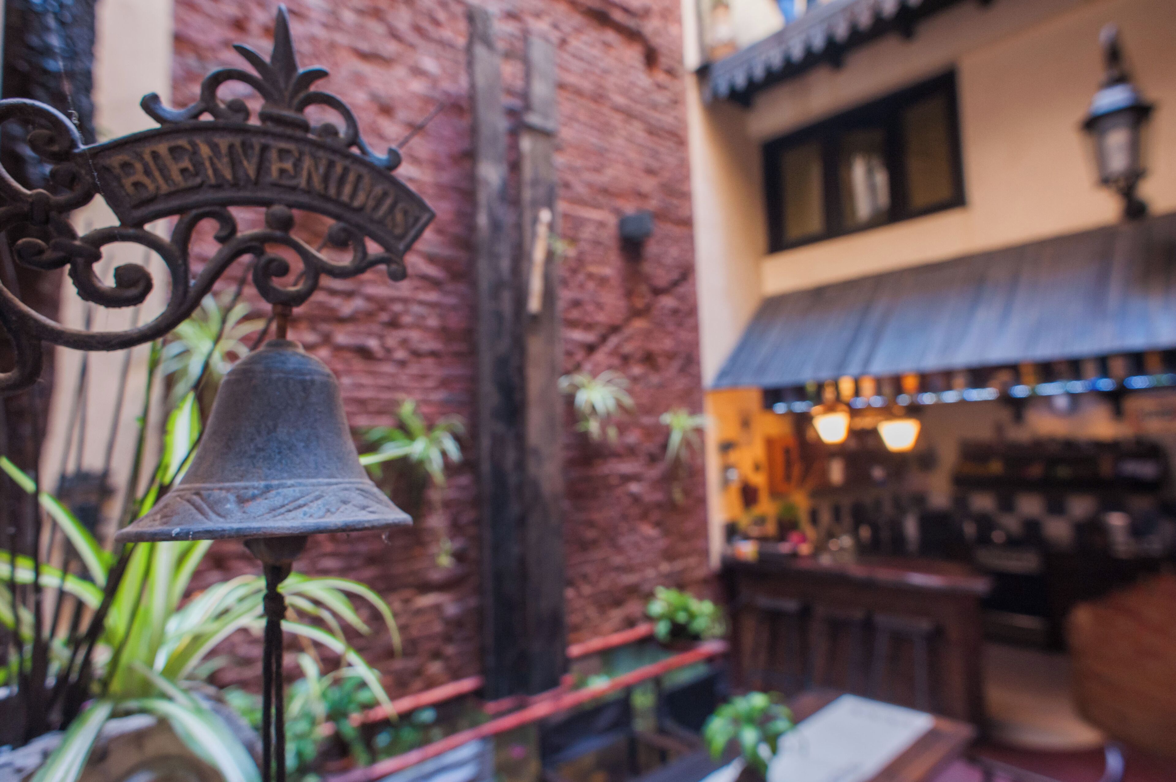 Una campana con el cartel "BIENVENIDOS", una pared de ladrillo y una zona de bar con plantas.