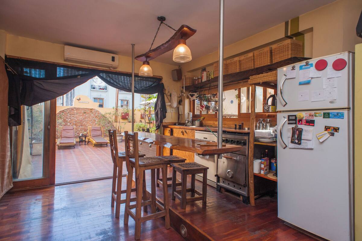 A compact kitchen with wooden furniture, a refrigerator, and a view through a glass door to an outdoor area with a brick wall and lounge chairs.