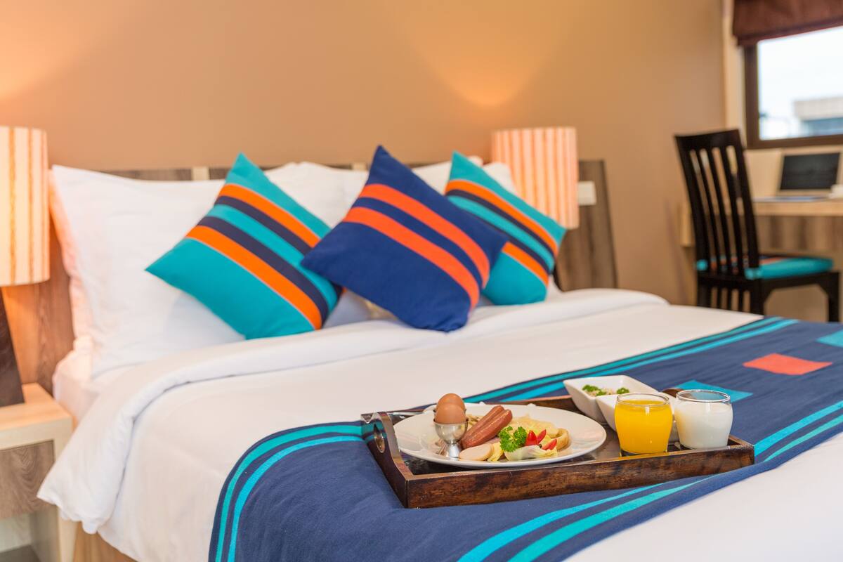 A neatly made bed with striped pillows, a tray with breakfast including eggs, sausage, and juice, and a laptop on a desk in the background.
