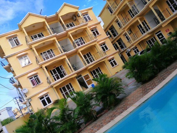 A multi-story yellow building with balconies, a swimming pool, and surrounding greenery.