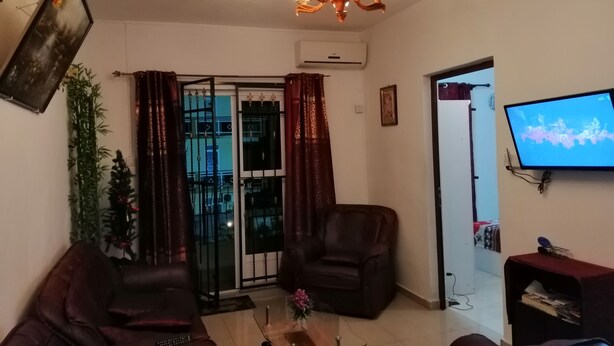 Un salon comprenant un canapé, un fauteuil et une table basse. Un téléviseur est fixé au mur et un lustre est suspendu au plafond.