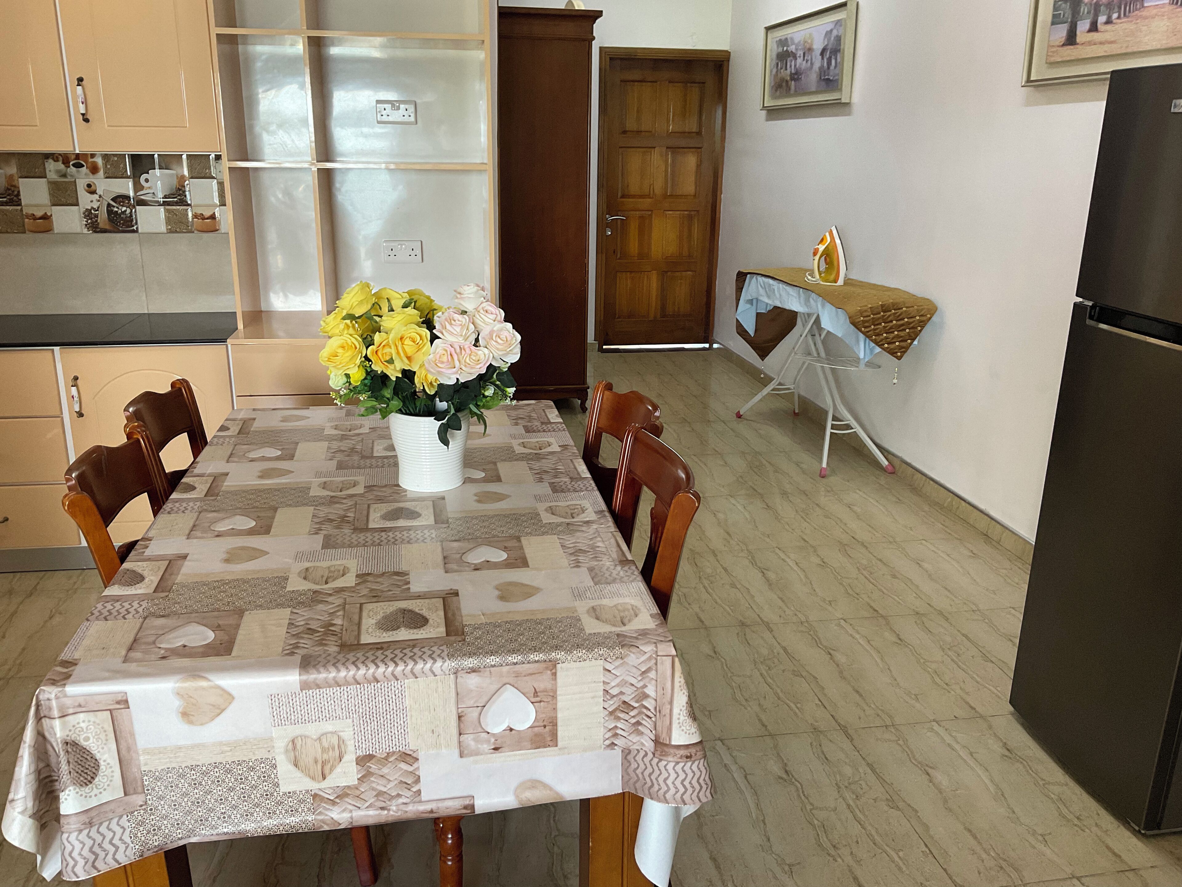 Un comedor con una mesa cubierta por un mantel estampado, rodeada de sillas de madera. Un jarrón con flores amarillas y rosas está sobre la mesa.