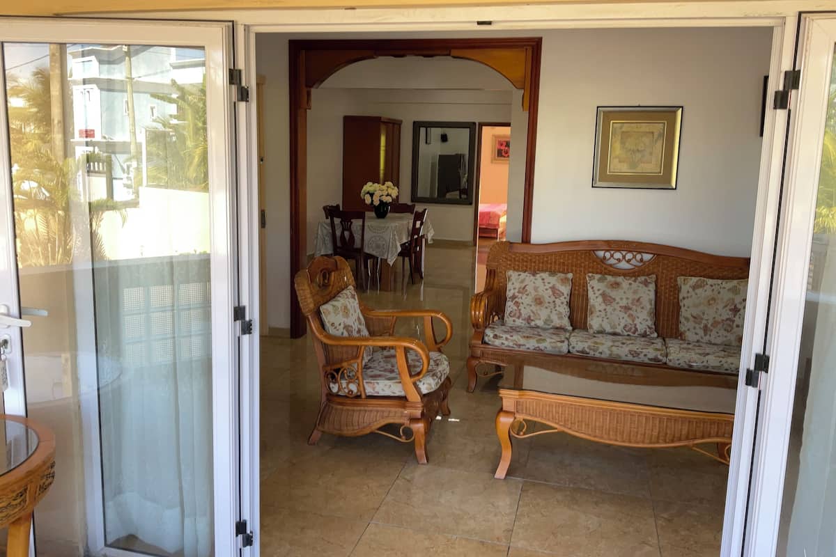 A living room with wicker furniture, a dining area with a table and chairs, and a kitchen in the background.