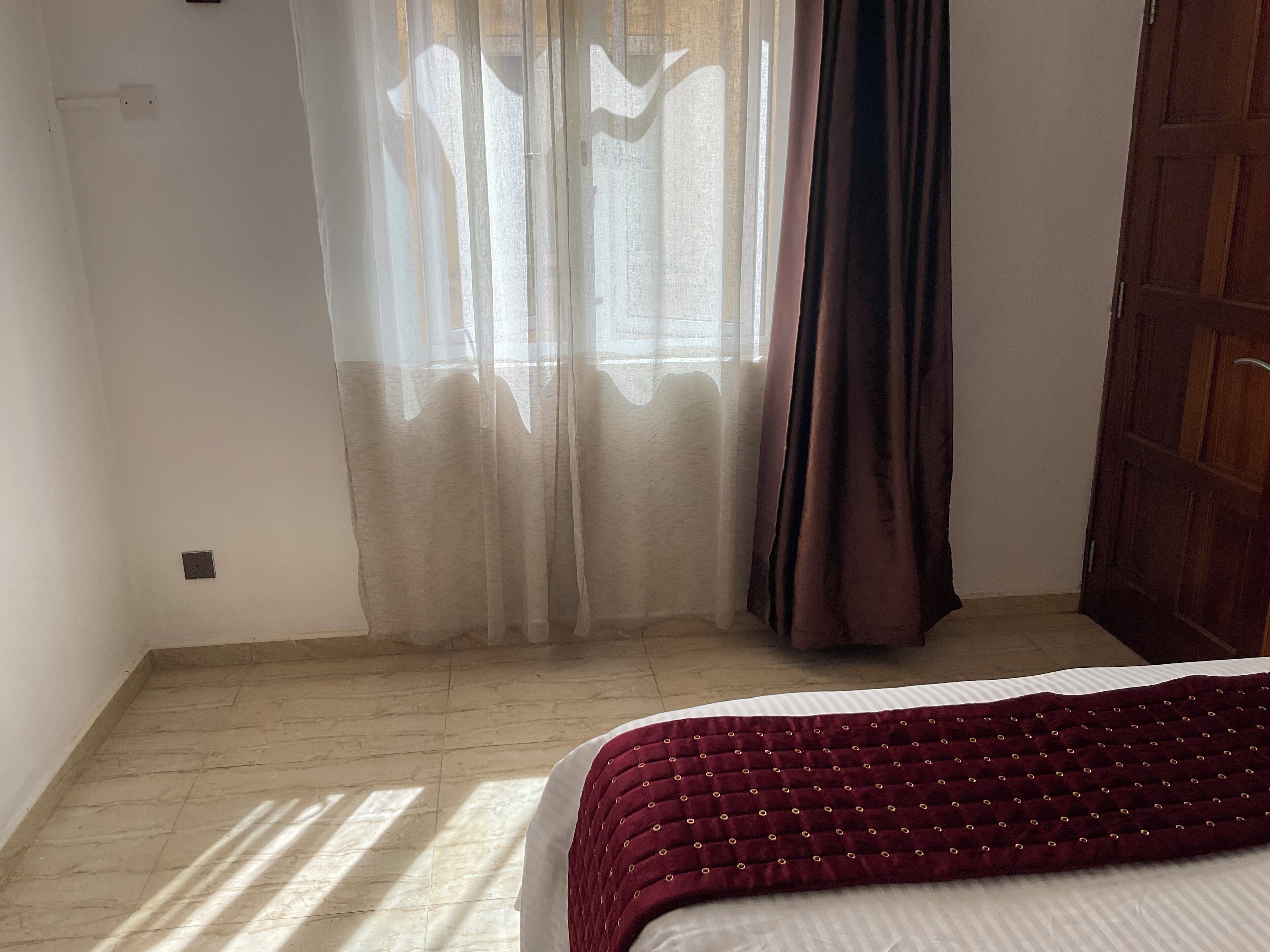 Une chambre à coucher avec un lit, une fenêtre avec des rideaux légers, un rideau plus épais et une porte en bois.