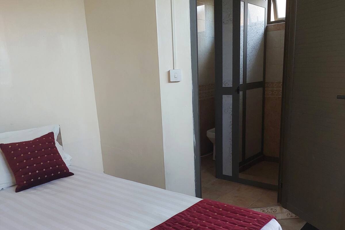 A neatly made bed with a white and red bedspread, a single red pillow, and a light switch on the wall.