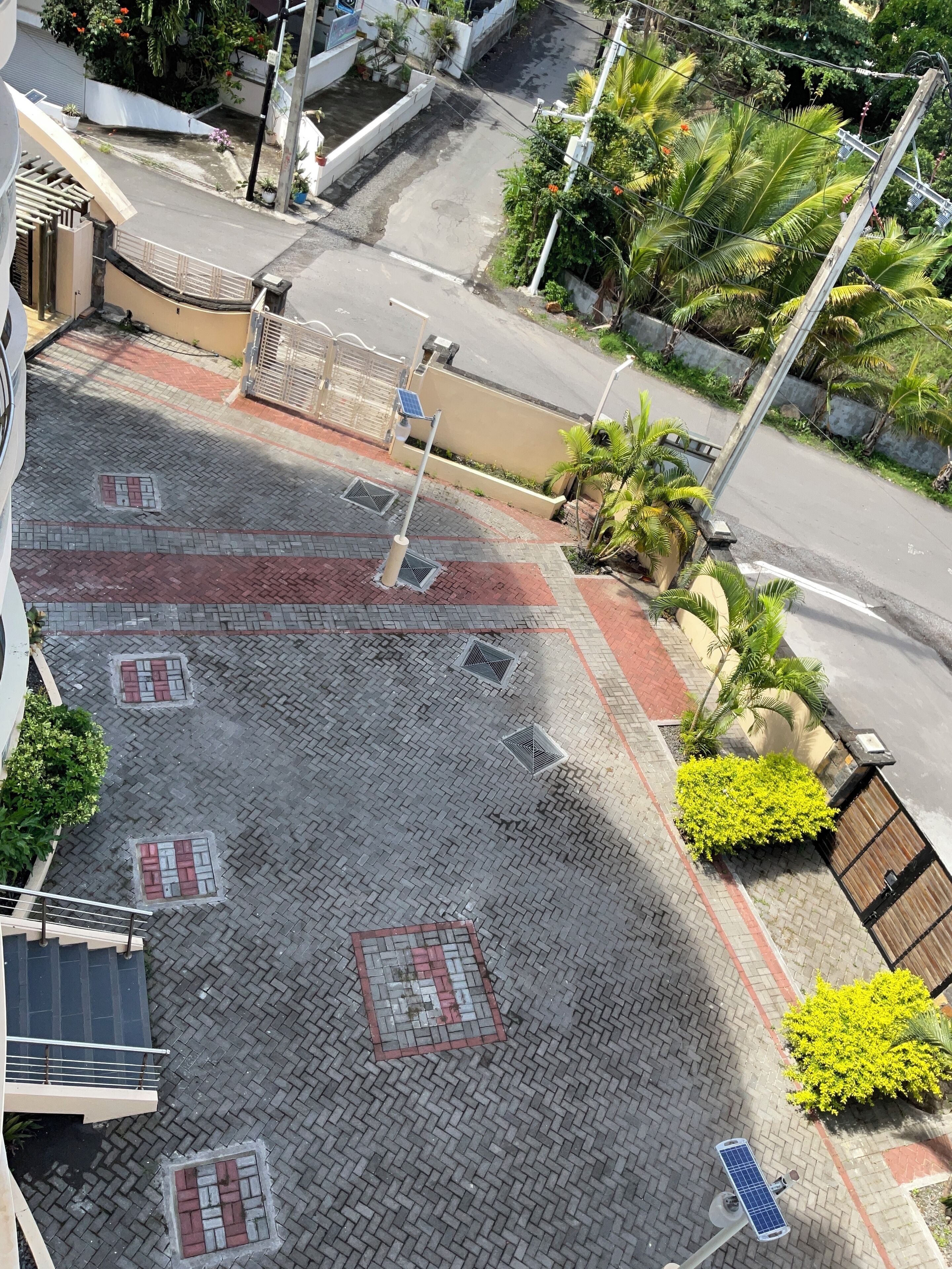 Une cour dallée, dotée d’une entrée fermée par un portail, de lampadaires solaires et de plantes en pot.