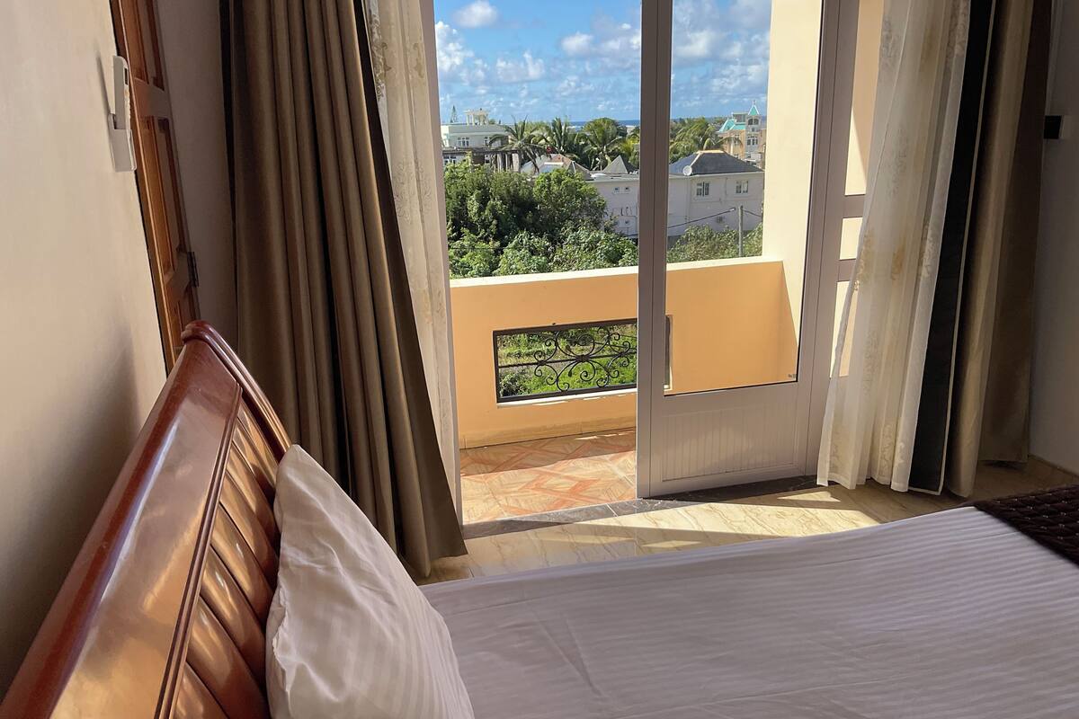 A hotel room with a bed, curtains, and a view of buildings and greenery through the window.