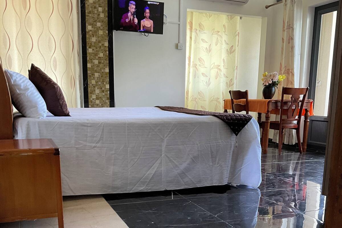 Quarto de hotel com cama, televisão fixada na parede, área de jantar com mesa e cadeiras, e piso revestido com azulejos.
