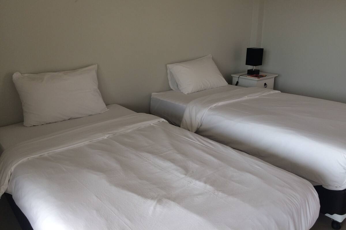 Two single beds with white linens in a room with plain walls and a small bedside table with a lamp.