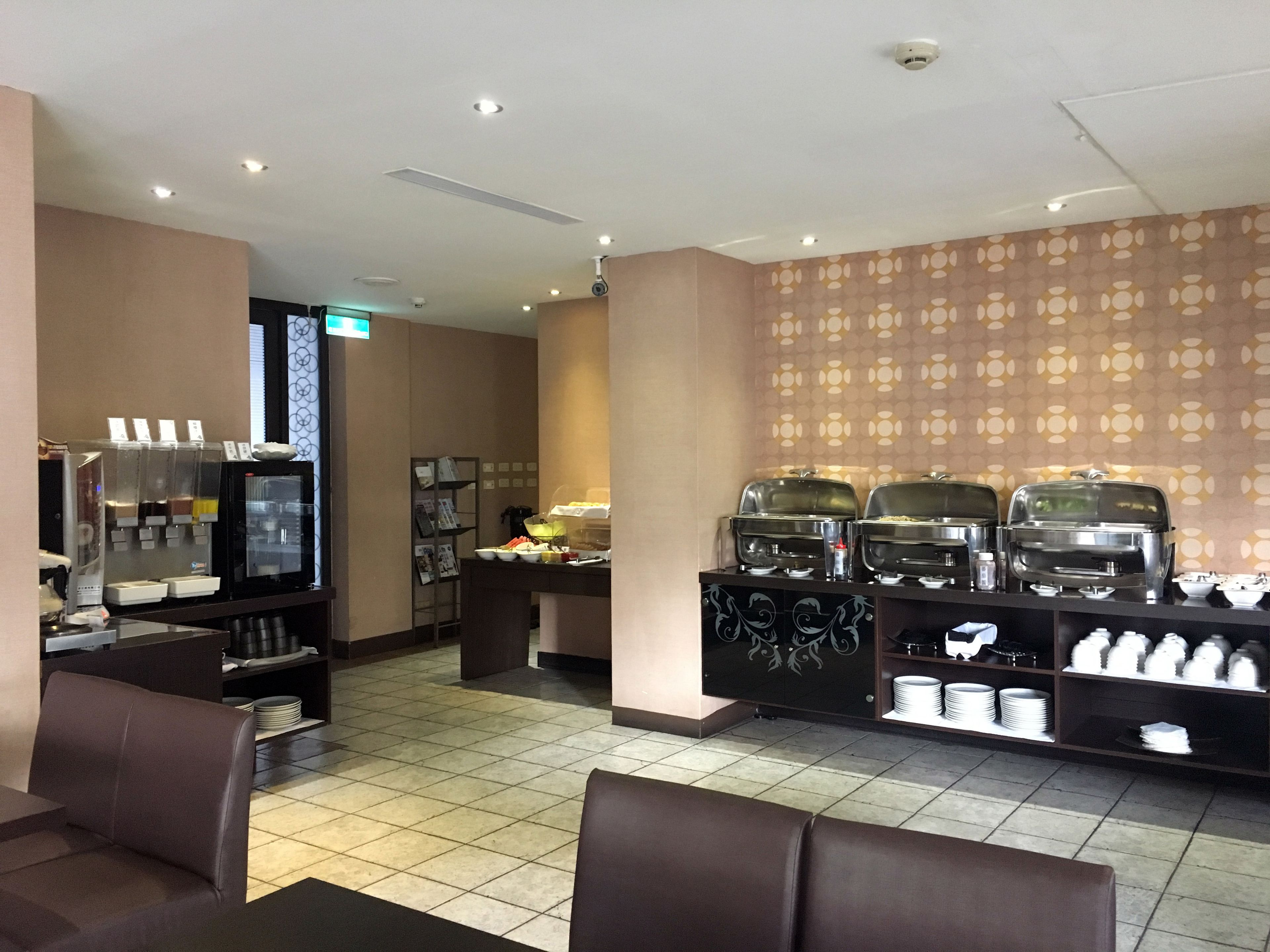 A hotel breakfast area with a coffee machine, buffet with hot food, seating area, and a patterned wall.