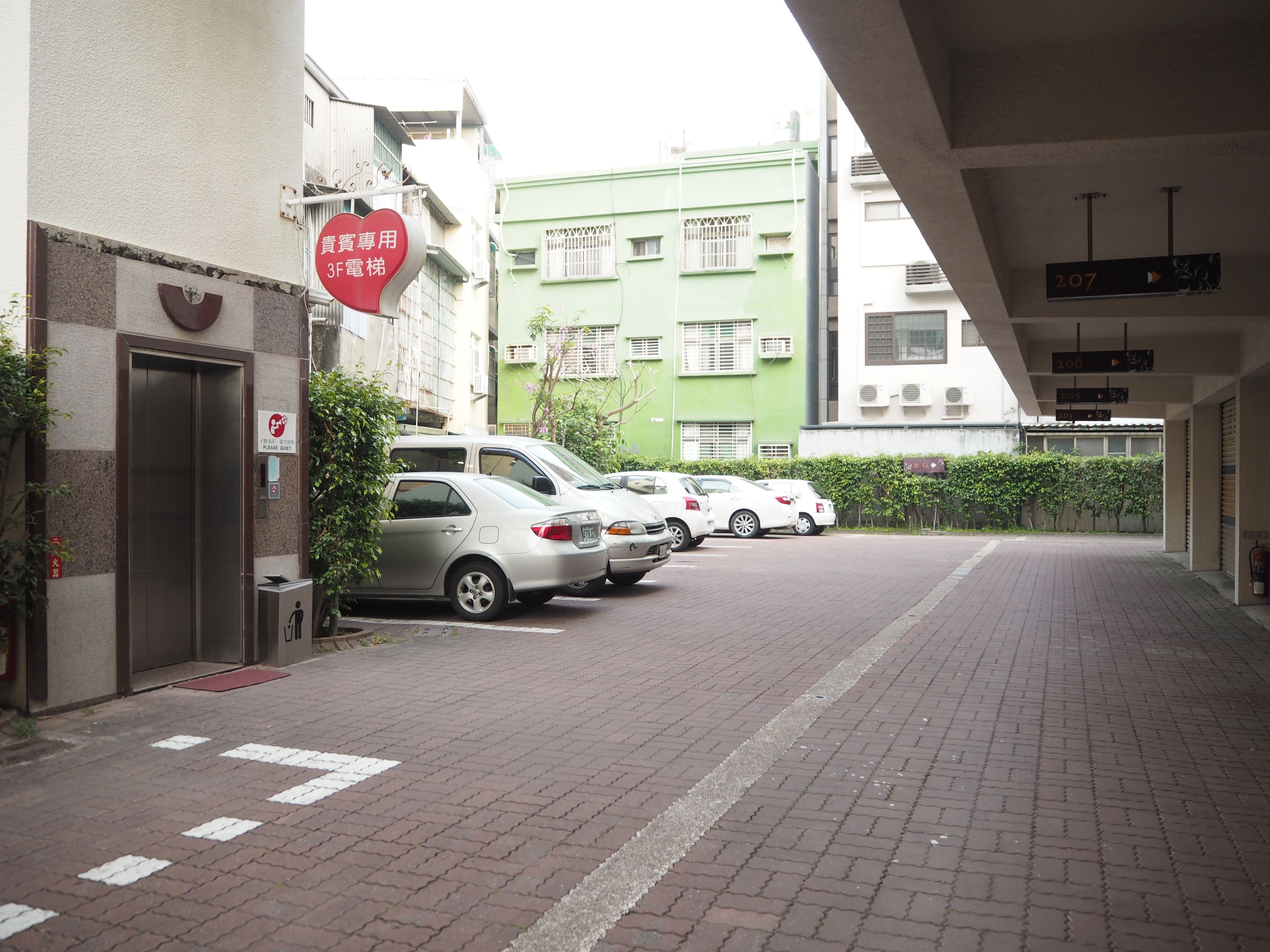 A parking area with cars parked along a brick-paved walkway, a building entrance with a red sign, and a green building in the background.