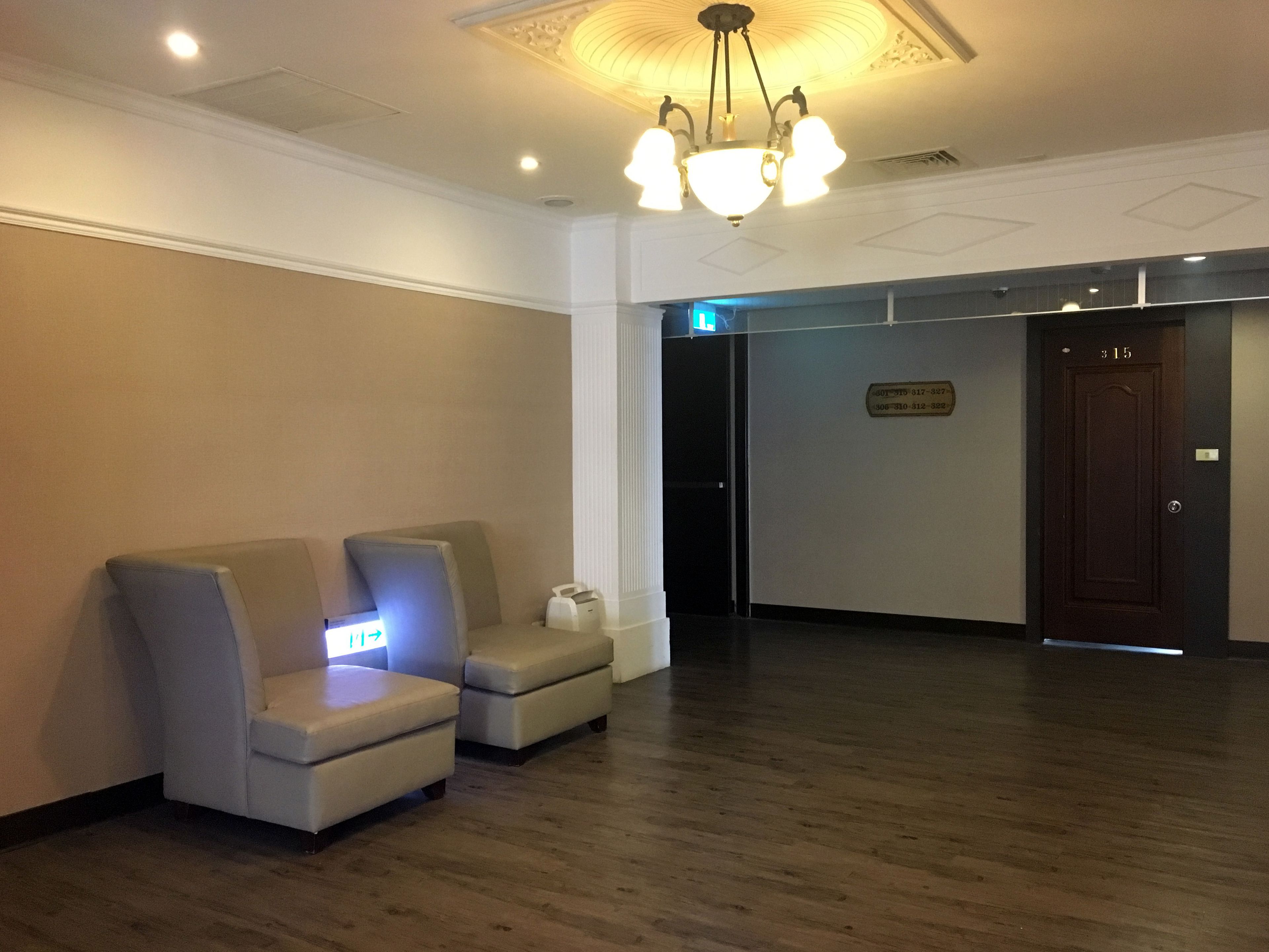 A hotel lobby with a reception desk, two armchairs, a wooden floor, and a chandelier.