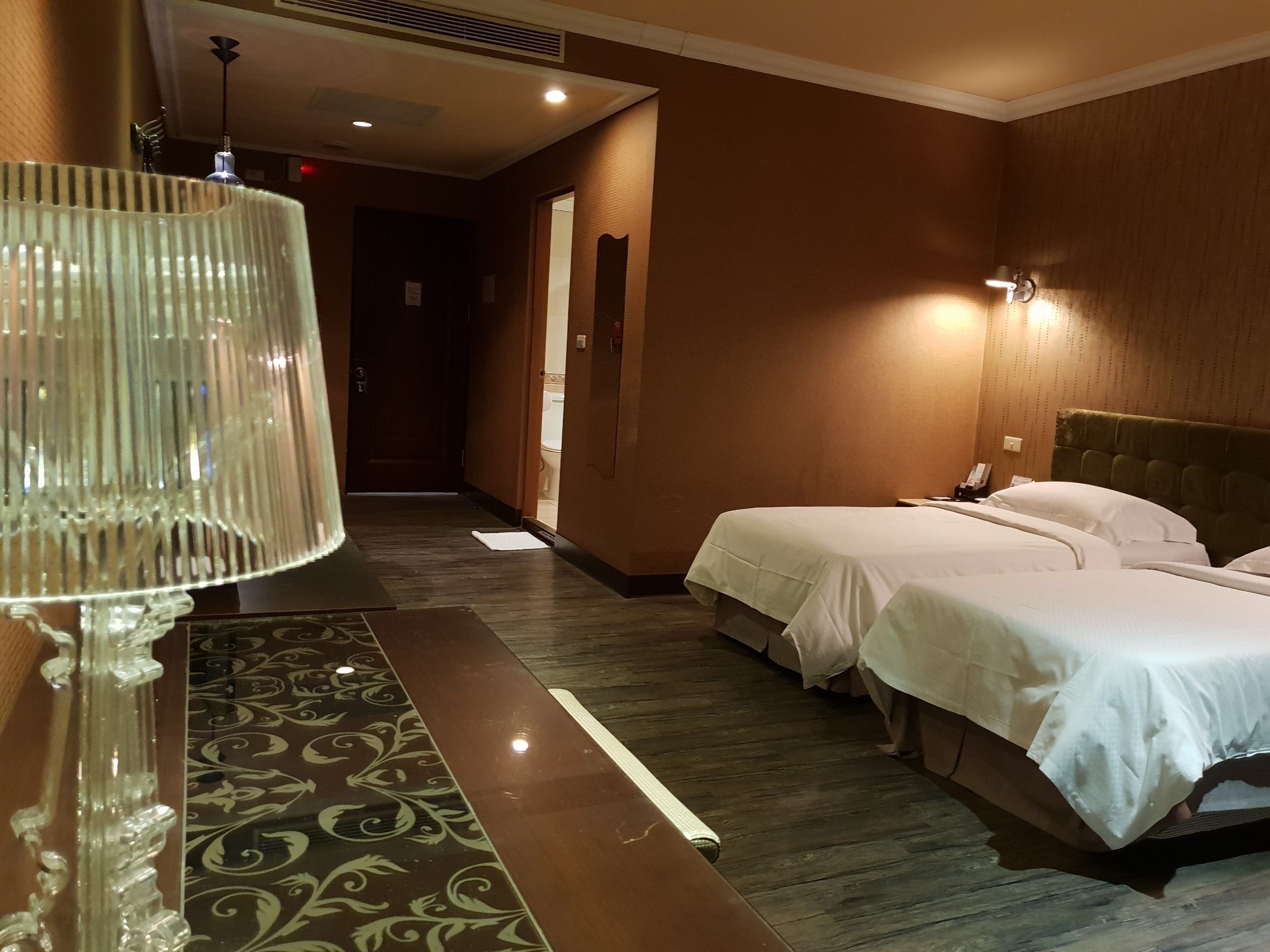 A hotel room with two beds, a bedside table, a lamp, and a patterned rug.